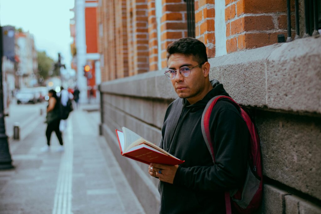 A young man reading outside against a brick wall in a city setting.