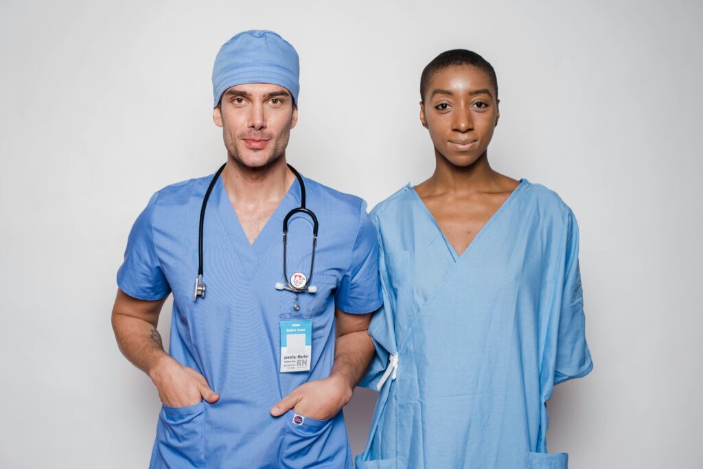 Positive male doctor in blue uniform with hands in pockets smiling and looking at camera while standing near nurse holding hands behind back in hospital