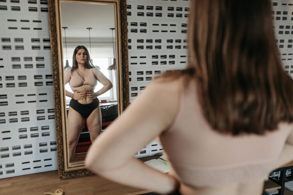 Woman in underwear looking at her reflection in a mirror, contemplating body image.