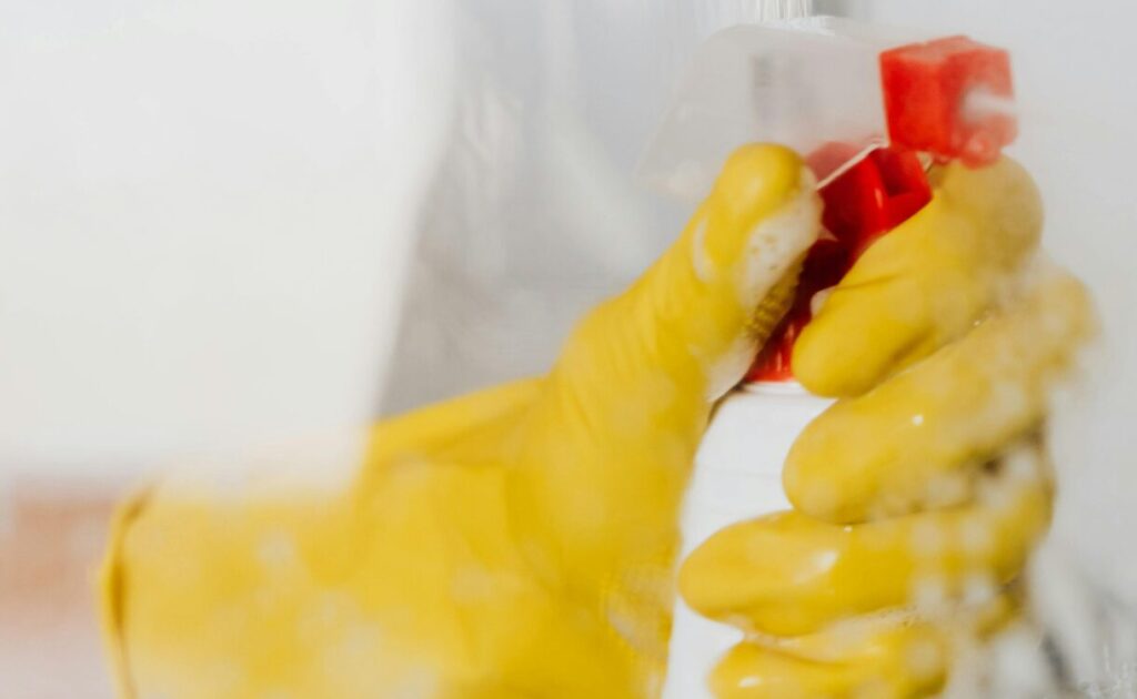 Crop unrecognizable person in yellow latex protective glove spraying detergent from bottle with plastic dispenser at glass of shower cabin