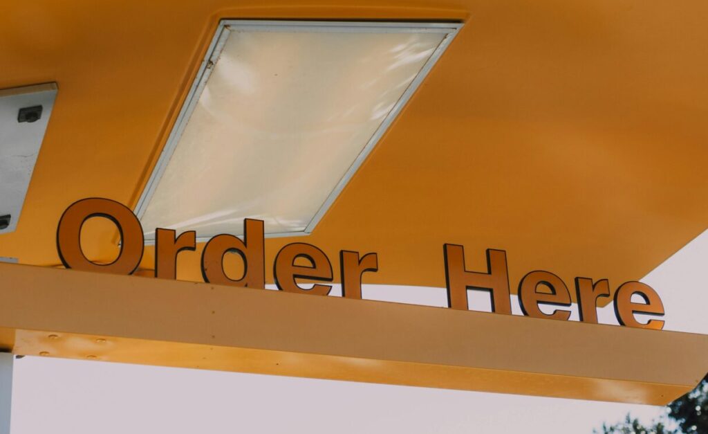A modern drive-thru order sign with yellow canopy under a clear sky, surrounded by trees.